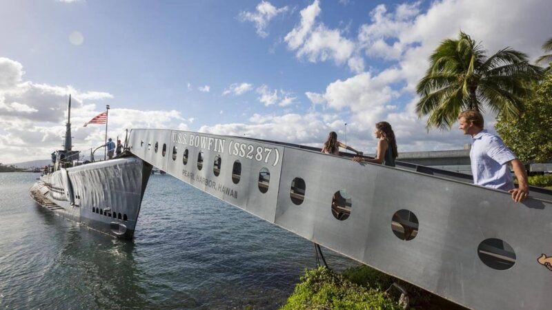 Visiteurs descendant une passerelle métallique pour embarquer à bord du sous-marin USS Bowfin amarré dans la baie, avec un drapeau américain flottant au soleil.