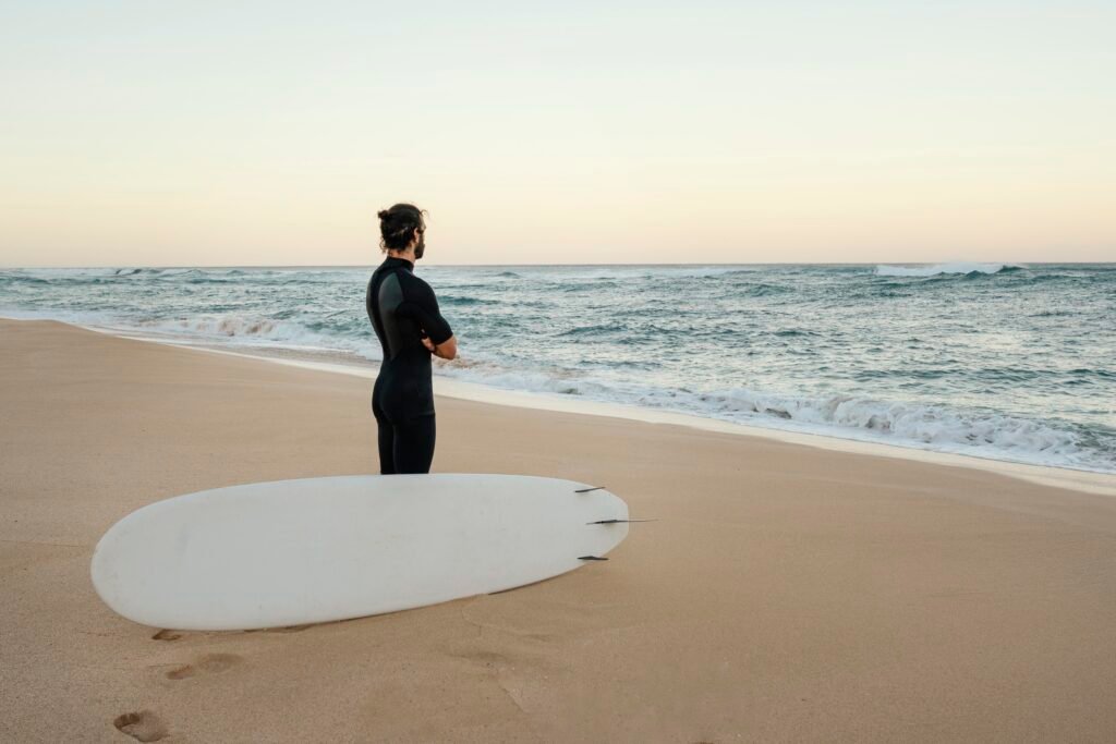🏄 Un surfeur en combinaison noire se tenant sur une plage de sable doré, contemplant l’océan avec sa planche posée à ses pieds, prêt à affronter les vagues légendaires d’Hawaï.