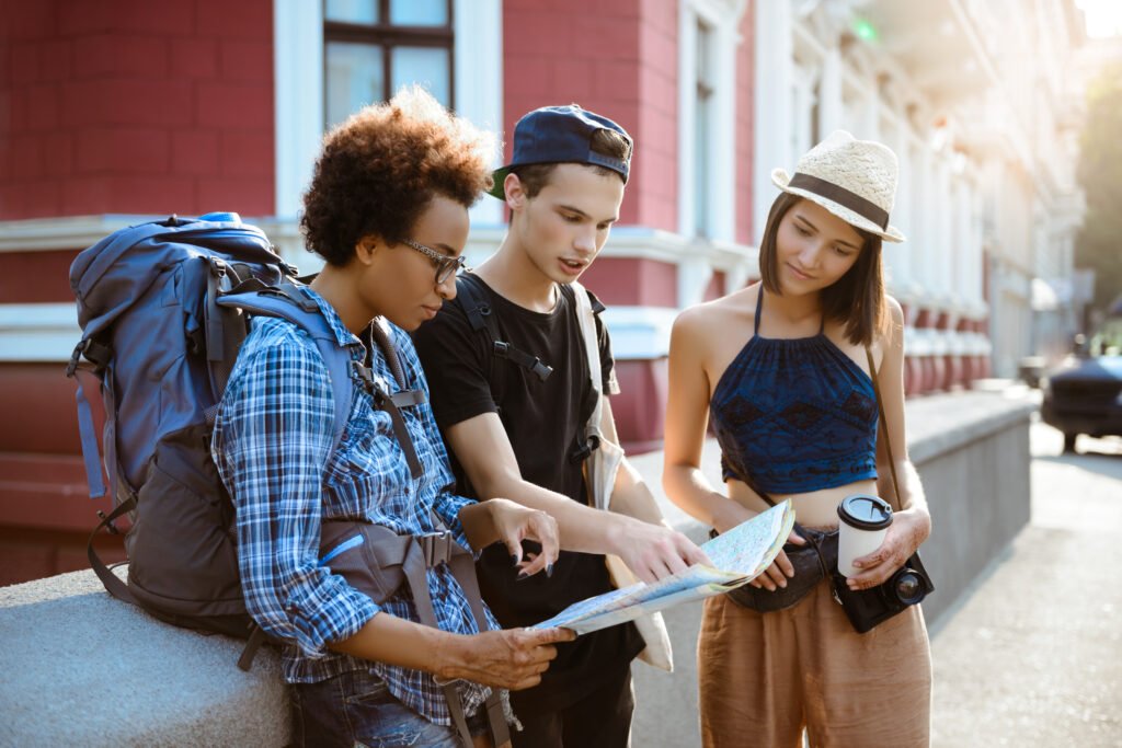 👉 Trois jeunes voyageurs explorant une ville avec une carte, vêtus de tenues décontractées et portant des sacs à dos, en pleine discussion sous un soleil éclatant.