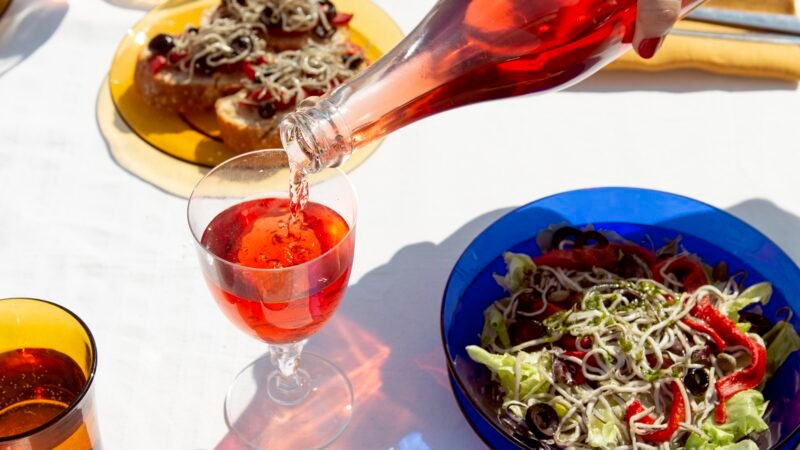 Table dressée en extérieur avec des assiettes colorées de salades et de plats de fruits de mer, tandis qu’une main verse un verre de boisson rouge.