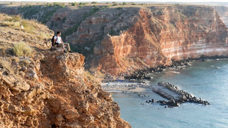 Un homme assis au bord d’une falaise rocheuse, regardant la côte escarpée et la mer en contrebas.