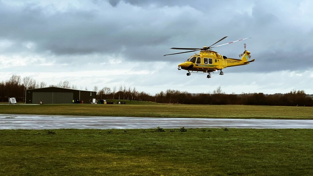 Hélicoptère jaune Safari Helicopters AW169 volant à basse altitude au-dessus d’une piste herbeuse, sous un ciel nuageux.