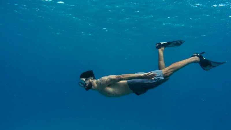 Homme en apnée portant un masque et des palmes nageant sous l’eau bleue.