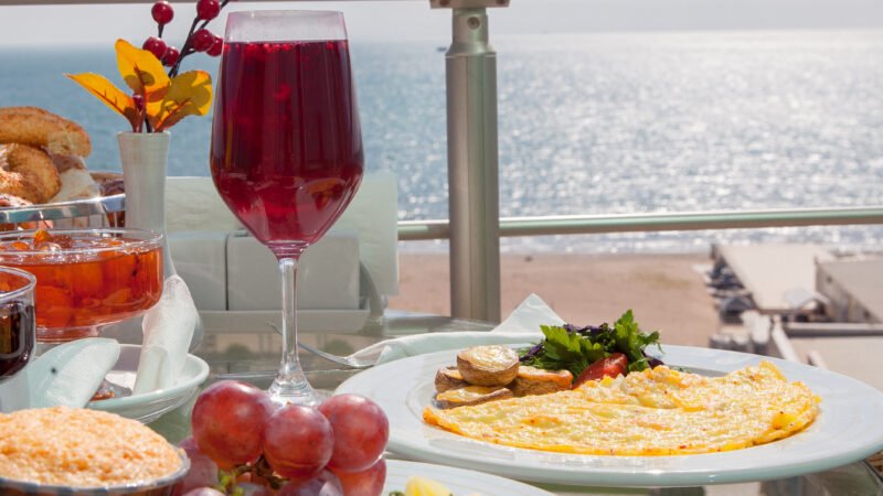 Table de petit-déjeuner dressée sur un balcon face à l’océan, comprenant une assiette d’omelette, un verre de jus rouge, des raisins et des pâtisseries.