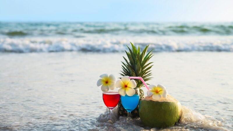 Deux cocktails colorés (rouge et bleu) décorés de fleurs de frangipanier, un ananas et une noix de coco garnie d’une paille, posés sur le sable humide à marée montante.