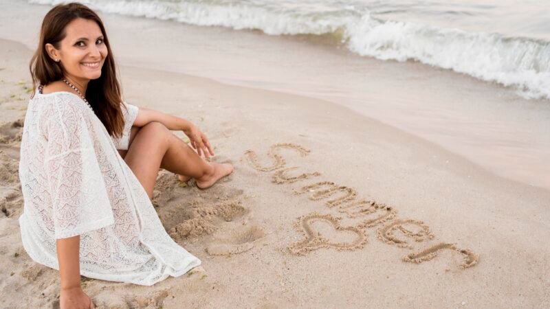 Femme souriante assise sur le sable, face à l’océan, avec le mot 'Summer' écrit sur la plage