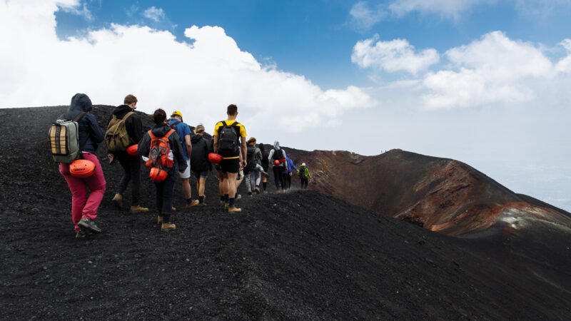 "Randonneurs en file indienne gravissant un sentier volcanique noir sous un ciel bleu, illustrant l’exploration du volcan Kilauea à Hawaï"