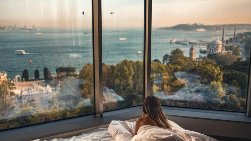 Femme allongée dans un lit face à une grande baie vitrée avec vue panoramique sur la mer