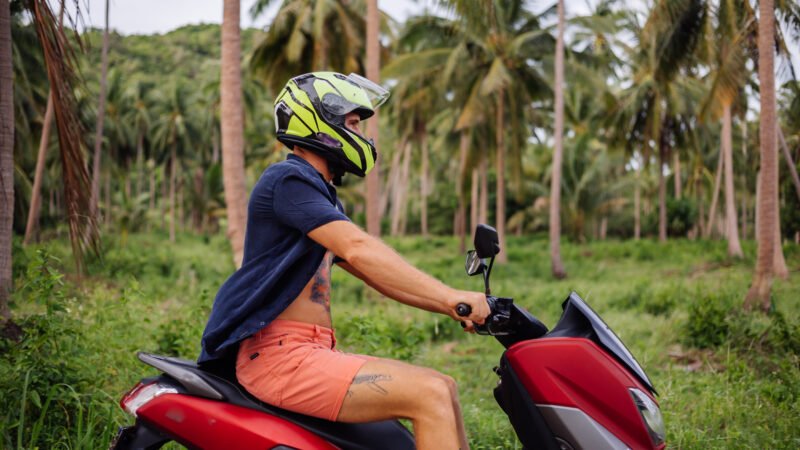 Homme tatoué portant un casque de moto, conduisant un scooter rouge à travers une zone tropicale verdoyante à Hawaï.