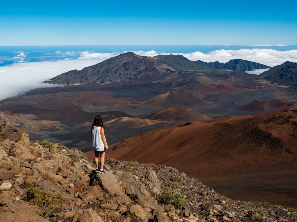 Femme debout sur un rocher au sommet du cratère Haleakalā à Hawaii, contemplant un paysage volcanique aride sous un ciel bleu et une mer de nuages.