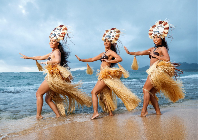 Trois danseuses polynésiennes en tenue traditionnelle effectuent une danse sur une plage de sable à Hawaï, face à l’océan.