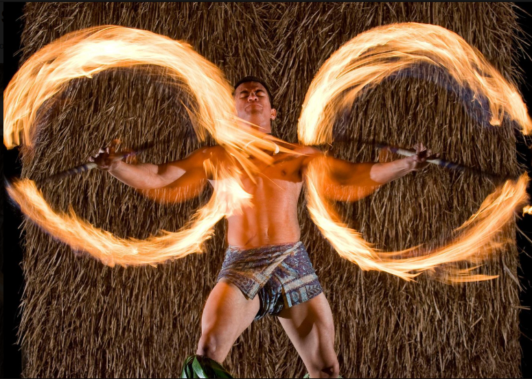 Danseur polynésien exécutant une danse de feu spectaculaire avec deux bâtons enflammés, sur fond de paille.