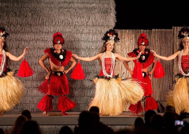 Groupe de danseurs polynésiens vêtus de costumes rouges et naturels, exécutant une danse traditionnelle sur scène pendant un luau, devant un public attentif.