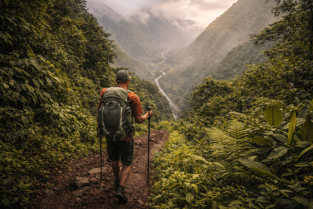 Randonneur descendant un sentier escarpé au milieu d’une jungle tropicale dense dans la vallée de Waipiʻo, avec vue sur la vallée brumeuse en contrebas.